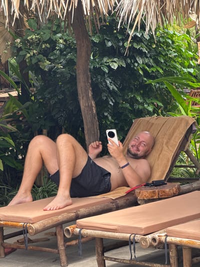 A bald, Caucasian man with a beard is reclining on a brown towel draped over a bamboo lounge chair. He is wearing black swim trunks and is holding a white smartphone in his right hand, looking at it. He is smiling. The lounge chair is made of bamboo and has a brown cushion. Next to the chair is a small wooden table with a black object and a red cord on it. The man is outdoors, with lush green foliage behind him and a thatched roof overhead. The scene appears to be in a tropical or subtropical environment. The location is identified as Nam Hoa Lư Ward, Vietnam.