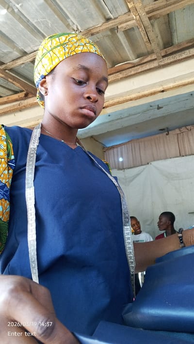 The image depicts a young Black female, likely engaged in tailoring, in the city of Gama, Nigeria. The timestamp in the lower left indicates 2026/03/19 14:37. The primary subject is positioned in the foreground, looking downwards with a focused expression. She wears a yellow headwrap with a green and black pattern, a dark blue V-neck top, and sleeves featuring patterned fabric. A white and black measuring tape hangs around her neck. Her left hand is partially visible in the bottom left, manipulating dark blue fabric. In the mid-ground, two young Black males are partially visible to the right. One male, on the left, wears a white t-shirt and looks to his right. The other male, on the right, wears a dark shirt and looks to his left, appearing to be in conversation. The background reveals a corrugated metal roof supported by exposed wooden beams and a light-colored wall or partition.