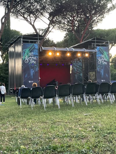 The image shows an outdoor concert stage in Rome, Italy at the location (latitude: 41.873, longitude: 12.494). The stage is set up in a grassy area, surrounded by trees. The stage is adorned with black banners that read "Casa Del Jazz Summer Time" and is lit by overhead lights. There are rows of chairs in front of the stage, where people are sitting and waiting for the performance to begin. One person stands near the edge of the frame, and a few people can be seen standing or sitting on the stage. A black grand piano is partially visible on the stage. The sun is setting and casting a golden light on the scene.