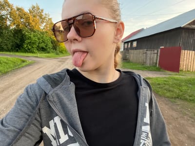 A young person, wearing large brown sunglasses and a grey zip-up hoodie over a black t-shirt, sticks their tongue out towards the camera. They have their hair pulled back and are wearing a small earring. The setting appears to be a rural area, possibly in Borodulino, Russia, with a dirt road, green grass, trees, and a wooden building with a red roof in the background. The sky is clear with a hint of sunset colors. The person is in the foreground, with the environment taking up the rest of the frame.