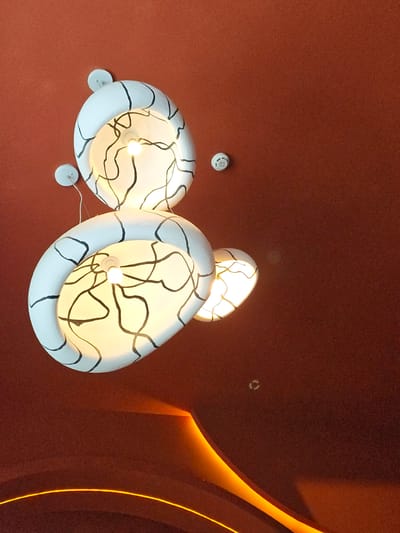 This is a low-angle shot of a modern ceiling with decorative lighting fixtures and an embedded orange light strip. The ceiling itself is a textured, dark reddish-brown color.
Dominating the upper left and center are two large, sculptural pendant lights. These lights are white with black, abstract, organic lines drawn on their interior surfaces. They appear to be made of a molded material. Each light has a visible bulb glowing warmly inside. Smaller, circular white ceiling fixtures are also visible, attached to the ceiling near the pendant lights, one with a visible wire extending downwards. To the right of the upper pendant light, a small, white circular object, possibly a smoke detector, is mounted on the ceiling.
In the lower half of the image, an embedded light strip glows with a vibrant orange hue, creating an abstract curved shape across the ceiling. This light strip suggests an architectural feature or accent lighting.
The overall impression is one of contemporary interior design, possibly in a commercial space or a well-designed residential interior. The lighting design is a focal point of the scene.
The provided context indicates this image is from Ashgabat, Turkmenistan. While the image itself does not contain explicit geographical markers, it suggests an indoor environment with modern architectural and design elements that could be found in various contemporary buildings within a city like Ashgabat. Without further information or visual cues within the image, it is not possible to pinpoint a specific location within Ashgabat.
