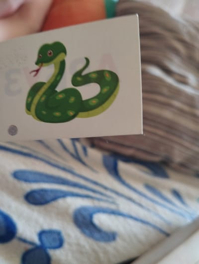 The image shows a close-up of a card with a cartoon snake on it. The card is white with a pale grey border on the left side. The snake is green with yellow accents and has red eyes and a forked red tongue. There is a faint grey number '3' to the left of the snake. In the lower part of the image, there is a white fabric with blue decorative elements, possibly a bedspread or blanket. A person's arm is visible in the upper left corner of the image, out of focus and with a reddish-orange sleeve. The image is not taken in the city of Kingisepp, Russia.
