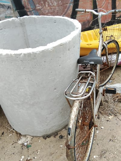 The image depicts a rusty, weathered bicycle leaning against a large, rough, concrete cylinder. The bicycle has a basket attached to the handlebars and a rear rack. The seat of the bicycle has the brand name "AMASA" visible. Behind the bicycle, a bright yellow object, possibly a kayak or a large float, is visible. The background shows a wall with graffiti. The ground appears to be a mix of sand and debris, with some dry grass and small stones. The overall scene suggests an outdoor urban or coastal environment, potentially in Phan Thiết, Vietnam, where such items might be found near recreational areas or storage spaces. There are no people or animals in the image.