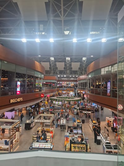 The image shows the interior of a large, modern airport terminal, likely in Pendik, Turkey, given the presence of "BIGCHEFS" branding, a popular Turkish restaurant chain. The scene is viewed from an elevated perspective, looking down onto multiple levels of shops and restaurants.
The architecture features a high ceiling with exposed structural elements and bright lighting. Wooden accents and large glass windows are prominent throughout the terminal. On the upper level, the "BIGCHEFS" restaurant is visible with diners seated inside. Below, various retail outlets are open, including shops for luxury brands like Bvlgari, Tom Ford, Dior, Guerlain, Chanel, and Balmain.
There are numerous people moving through the terminal, browsing shops, dining, and conversing. Some are carrying luggage, indicating travel. Employees are visible working at the retail counters. The overall atmosphere suggests a busy and commercial environment. The image captures a typical airport scene with a focus on retail and dining facilities.