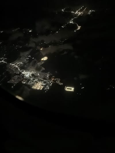 This aerial night view shows a section of Santa Ana, California, United States, illuminated by city lights. The scene is dominated by darkness, with scattered clusters of lights representing urban development. Prominent features include a densely lit rectangular area with a grid-like pattern, possibly industrial or commercial buildings, and another brightly lit rectangular structure, which may be a sports stadium or a large complex. Surrounding these areas are smaller, less concentrated groups of lights, indicating residential neighborhoods or less developed zones. Faint cloud cover is visible, softening the glow of the lights in some areas. The perspective is from a high vantage point, suggesting the image was captured from an airplane or drone.