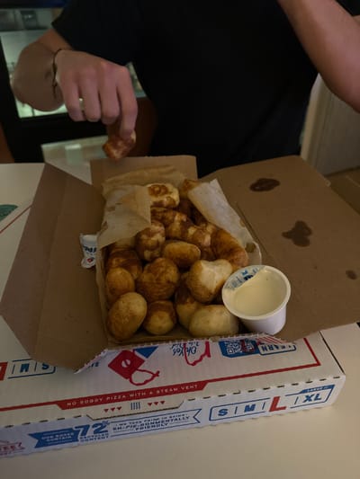 The image depicts an indoor scene, presumably in Boston, Massachusetts, United States, showcasing a Domino's Pizza box containing Cinnamon Bread Twists. A person, wearing a dark short-sleeved shirt and a dark bracelet on their left wrist, is visible, with their right hand actively retrieving one of the golden-brown, cinnamon-sugar-coated bread twists from the box.
The open, white cardboard Domino's box, displaying red and blue branding, rests on a light-colored, flat surface. Visible text on the box includes "NO SOGGY PIZZA WITH A STEAM VENT" and "OUR BOXES CONTAIN 72% RECYCLED CONTENT," along with size indicators (S, M, L, XL). The inside flap of the box contains several irregular dark stains. Inside the box, a substantial quantity of Cinnamon Bread Twists is piled within a brown paper liner. A small white plastic cup of white icing, with its foil lid partially peeled, is positioned among the bread twists. A smaller, mostly obscured white cup with blue branding is visible to the left. The background is dimly lit and indistinct.