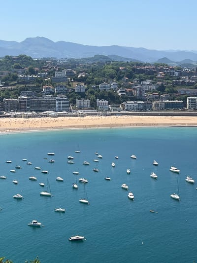 This is an elevated, wide-angle, medium-shot photograph capturing a panoramic view of La Concha Bay in San Sebastián, Spain, under a clear, bright blue sky. The scene is divided into three distinct horizontal bands: the azure blue sea in the foreground and middle ground, a golden sand beach with numerous people in the middle ground, and a cityscape with buildings nestled among green hills and mountains in the background.

The sea is dotted with a multitude of white boats, ranging from small motorboats to several sailboats, anchored or slowly moving across the water. Some boats appear to be yachts, while others are simpler vessels. A few figures can be seen in the water, either swimming or on small inflatable devices.

The beach is crowded with sunbathers, and the sand is a warm, light brown color. Numerous umbrellas are visible, creating a mosaic of colors on the beach. People are spread out, with some closer to the water's edge and others further back.

The cityscape in the background features a dense collection of buildings with varying architectural styles, predominantly white and light-colored facades. These buildings are situated on a hillside that is densely covered with lush green trees and vegetation. Beyond the city, a series of rolling mountains, also covered in vegetation, recedes into the distance. The overall impression is one of a vibrant, sun-drenched coastal city on a beautiful summer day.
