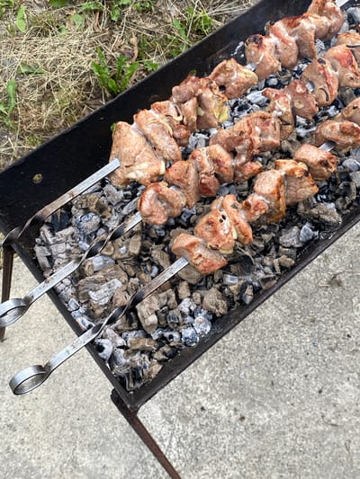 In this top-down, medium shot, several skewers of meat are being cooked over hot coals in a black metal brazier. The meat, cut into uniform cubes, appears to be pork and shows signs of browning and charring from the heat. Some pieces have visible marbling and fat. The skewers are made of metal and have twisted handles. They are arranged in a row across the brazier, with the meat pieces interspersed with what appear to be slices of onion, though this is difficult to confirm definitively.

The brazier sits on a surface of hot, grey coals, some of which are glowing. The coals are situated within the rectangular confines of the brazier, which shows signs of wear and tear. The brazier is placed on a textured grey concrete surface, suggesting an outdoor setting. To the left of the brazier, a patch of dry grass and green vegetation is visible, hinting at a rural or semi-rural location. A metal rod, likely part of the brazier's stand or a tool, extends out of frame to the right.

The lighting suggests it is daytime, with no strong shadows indicating a specific time. The overall scene evokes a sense of outdoor cooking, likely a barbecue or a similar gathering. The focus is entirely on the food and the cooking process, with no people visible. The image is static, capturing a moment in time during the preparation of a meal. There is no visible text in the image.