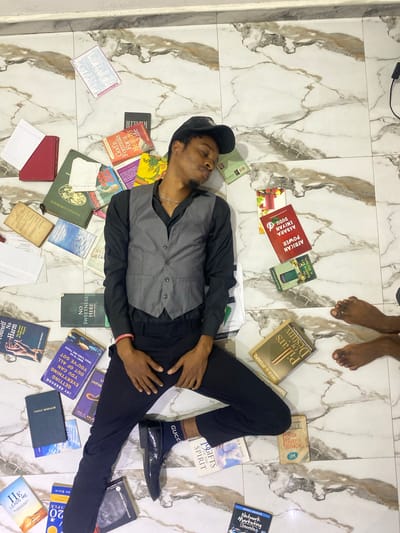 A man is lying on a marble-tiled floor in an indoor location. The environment is filled with books scattered around him. The man is wearing a grey vest over a black shirt, black pants, black shoes with Gucci socks, and a black cap. He is positioned diagonally across the frame, with his head near the top left and his feet towards the bottom right. The books surrounding him have various titles and colors. There is also a pair of bare feet visible near the top of the image. There is no indication of the city Joma, Ghana, in the image.