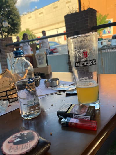 This is a close-up, eye-level, outdoor shot of a table at what appears to be a cafe or restaurant, likely located in the city of Yunus Emre, Turkey, as indicated by the "damla" water bottle which is a Turkish brand and the text on the cigarette packs are in Turkish. The foreground features a wooden table with several objects on it. To the left is a partially filled glass bottle of "damla" water. Next to it is a smaller glass containing clear liquid. Further to the right is a tall glass with a salt-rimmed edge, containing a yellow beverage, possibly beer, with the "BECK'S" logo visible. In front of the beer glass are two packs of cigarettes, one "Winston Slender Blue," and a red disposable lighter. There are also some scattered crumbs and a small ashtray. In the background, out of focus, there are people, brick structures, and some greenery, suggesting an urban outdoor setting. The lighting indicates it is daytime, likely late afternoon due to the warm light.