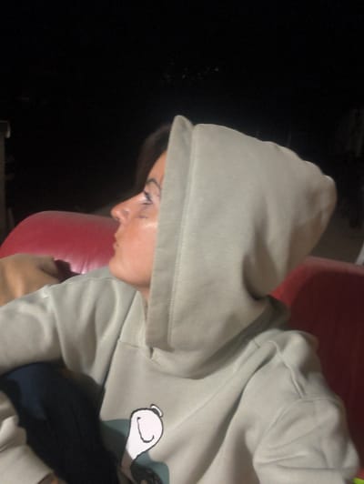 A young woman is depicted in a profile view, looking to the left of the frame. She has dark hair, a nose piercing, and glitter or makeup applied around her eye. She is wearing a light-colored hooded sweatshirt, appearing sage green or light grey, which features a cartoon character graphic on the front – a white head with black outline for eyes and ear, resembling a popular cartoon dog.
She appears to be seated or leaning back against a soft, red surface, likely a cushion or part of a chair, which fills the background immediately behind her. The rest of the background is very dark and indistinct, suggesting a nighttime setting or a dimly lit indoor space. The lighting is low, illuminating her face and the front of her sweatshirt. Her expression is neutral, possibly contemplative. There is no visible text in the image.