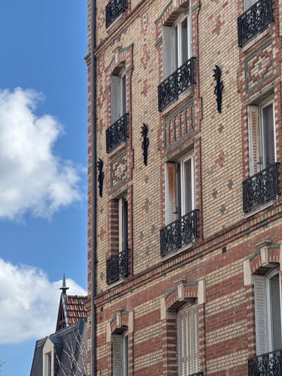 An image taken in Bois-Colombes, France, depicts the upper section of a multi-story building facade under a clear blue sky with scattered white clouds. The building features a brick exterior with alternating bands of light beige and red bricks, interspersed with teal-colored brick accents forming geometric patterns, notably around the window frames. Multiple rectangular windows are visible, each equipped with white frames and white louvered shutters; some shutters are fully open, some partially open, and some closed. Most windows are fronted by decorative black wrought iron balconettes or railings. Black, stylized vertical ornaments are affixed to the brickwork between window columns. A vertical drainpipe runs along the left edge of the visible facade. To the lower left, a portion of a contrasting dark-tiled roof from an adjacent or lower building is visible, topped with a spired finial. No individuals or specific actions are discernible.