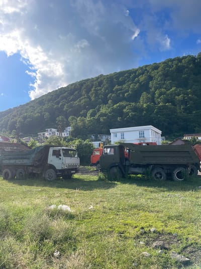 The image depicts an outdoor scene on a sunny day in Baranovka, Russia. In the foreground, two large dump trucks are parked on a grassy area. The truck on the left is lighter in color, while the one on the right is dark green. Behind the trucks, a densely forested green hillside dominates the background, with a few white buildings visible amidst the trees. The sky above is a mix of blue and clouds, with sunlight casting bright highlights. The environment appears to be a somewhat undeveloped area, possibly on the outskirts of a settlement, given the presence of the industrial vehicles and the natural landscape. There are no people visible in the image.