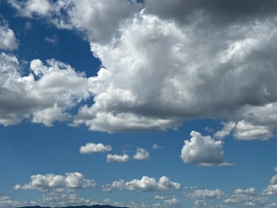 A wide shot captures a dramatic sky filled with a variety of clouds over a silhouetted landscape. The sky is a vibrant blue, with fluffy white cumulus clouds scattered throughout. Larger, darker storm clouds dominate the upper portion of the frame, casting shadows and creating a sense of depth. In the distance, a hazy, dark line suggests mountains or hills. The scene is devoid of any people or man-made structures, offering a purely natural vista. The overall impression is one of natural beauty and the power of the elements.
