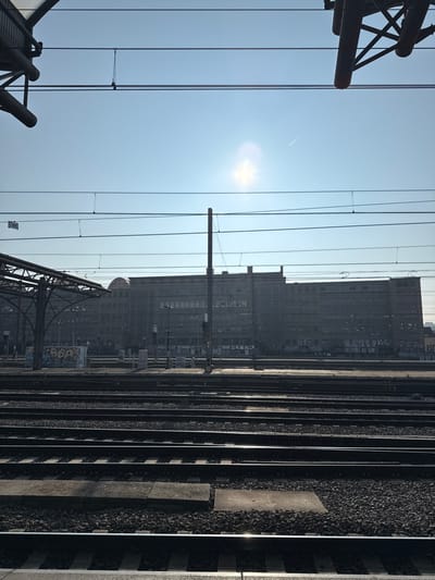 The image displays a wide view of a railway yard and platforms in Saint-Gilles - Sint-Gillis, Belgium, likely captured from a train or elevated station perspective. The scene is dominated by multiple parallel railway tracks, approximately ten sets, running horizontally across the lower two-thirds of the frame, composed of metal rails on gravel ballast and sleepers. Concrete platform sections are visible between some tracks.
In the midground, to the left, a dark, arched metal canopy structure covers a portion of a platform, adjacent to which is a graffiti-covered utility box displaying white and blue lettering. Several tall, dark utility poles are positioned between the tracks and the background building. Further back, additional platforms or concrete walls are visible, also marked with various textual graffiti.
The background is dominated by the Royal Belgian Institute of Natural Sciences building, a large, multi-story structure with a dark, gray facade and numerous windows, situated beyond the railway lines. A distinctive domed roof feature is prominent on the left side of this building. The lower sections of the building also display extensive graffiti.
The sky is clear and bright blue, featuring a prominent sun flare slightly right of center and a faint aircraft contrail in the upper right quadrant. Overhead, numerous electrical catenary lines crisscross the sky, supported by dark metal structures partially visible in the upper corners. Shadows are cast towards the right side of the frame, indicating the sun's position. No individuals are discernible within the immediate railway track or platform areas.