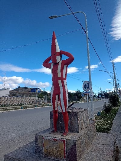 A red and white striped figure stands atop a stone pedestal, positioned beside a road with a 40 speed limit sign. The figure has a pointed head and appears to be wearing a form-fitting suit. Overhead, power lines run across a clear blue sky dotted with white clouds. The pedestal sits next to a sidewalk. In the background, there are houses, trees, and hills.