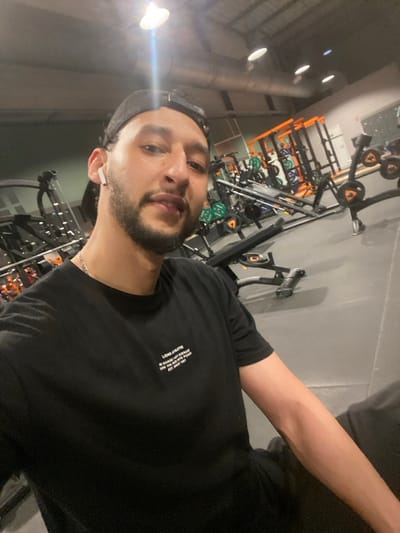 A male individual is depicted in a gym environment in Hœnheim, France. He has dark hair, a beard, and is wearing a black cap backward. A white wireless earbud is present in his left ear, and a silver chain is visible around his neck. He wears a black t-shirt with the white text "LIONS ATHLETIC IN SAVAGE / NOT AVERAGE TURN THE PAIN INTO POWER EST. 1997" printed on the chest. The man is looking directly at the camera, appearing to take a selfie. The background displays a fitness center with dark gray rubber flooring and various exercise equipment. This equipment includes weight racks with orange frames, barbells, weight plates (some green, some black with orange accents), and a flat bench. Overhead lighting illuminates the scene, with one bright light source producing a distinct lens flare near the top center. The ceiling is high, revealing exposed ductwork.