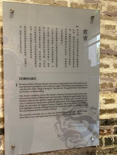 A clear acrylic or glass plaque is affixed to a rustic, light-colored brick wall, secured by four silver stand-offs. The plaque displays text in both Chinese and English, serving as a "Foreword" or "Forward" for an exhibition.

The English text, prominent on the right, explains that Bruce Lee's global recognition brought Chinese Kung Fu to the world's attention. It highlights his title as the "King of Kung Fu" and his "thoughtful fists." The text further describes the renovation of Lee Hoi-chuen's former residence (now Bruce Lee's ancestral home) and how the exhibition explores Bruce Lee's spiritual origin and growth as a world cultural icon. It also mentions showcasing the previous splendor of the Xiguan Mansion, the attraction of Lingnan culture (from the south of the Five Ridges), and the role of Kung Fu and Cantonese opera as a link between the motherland and overseas Chinese. The text concludes with a welcome to visitors to learn more about Bruce Lee and traditional Chinese culture. The Chinese text on the left conveys the same message.

A faint, stylized dragon-like watermark is visible on the right side of the plaque. The brick wall background suggests an old or historically preserved building, consistent with the description of an ancestral home and mansion. The overall setting appears to be an indoor exhibition space, likely within the Lee Family's ancestral home in Guangzhou, China.