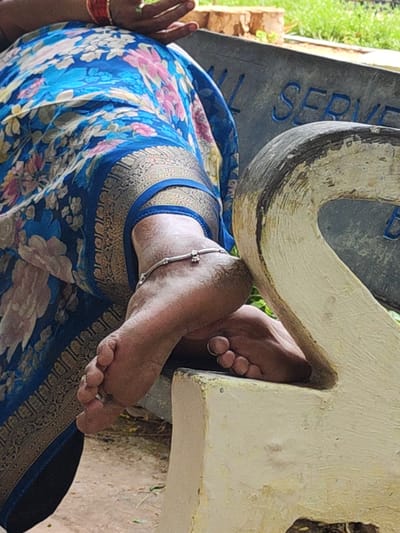 A close-up captures the feet of a person, seemingly female, adorned with a silver anklet, resting on a white painted structure, possibly part of a memorial stone. The individual is draped in a blue floral saree with a gold border. The location appears to be near a marble or granite plaque with letters partially visible. Given the context of Puttaparthi, India, this may be within the grounds of Prasanthi Nilayam or a related memorial site.