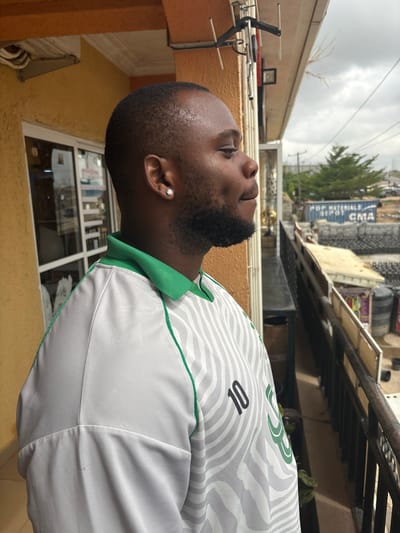 The image, taken in Lugbe, Nigeria, captures a profile view of a man looking upwards, set against an overcast sky and urban backdrop. The man has dark skin, short dark hair, and a neatly trimmed beard. He is wearing a single, sparkling diamond stud earring in his left ear.

He is dressed in a light-colored, possibly white or light grey, short-sleeved collared shirt with a green trim around the collar and sleeves. The shirt features a distinctive wavy pattern of darker grey lines. The number "10" is visible on the shirt, suggesting it might be a sports jersey or a uniform.

The man's gaze is directed towards the cloudy sky, which dominates the upper portion of the background. Below the sky, architectural elements are visible, including the edge of a building or balcony railing. To the left, an antenna-like structure is present on a textured, yellowish-brown wall, along with some wires and pipes. In the distance on the right, blurred buildings and green trees are visible. One prominent building in the background has text on its side, reading "POP MATERIALS DEPOT CMA". The overall setting appears to be an outdoor, likely urban or semi-urban environment, possibly from a balcony or rooftop during daytime with overcast weather.