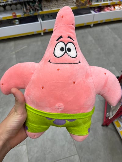 A close-up shot shows a pink, plush toy resembling Patrick Star from SpongeBob SquarePants. The toy is being held by a person's hand, partially visible on the left side of the frame. Patrick is wearing green shorts with purple polka dots. The background appears to be an aisle in a store, with shelves of merchandise visible. The flooring is gray and tiled. The overall lighting suggests an indoor retail environment. The scene does not provide any information about the city of Ivanivka, Ukraine.