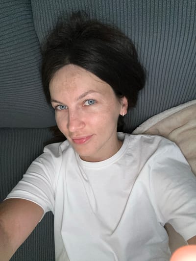 A woman with dark hair and blue eyes is lying on a textured grey couch. She is wearing a white t-shirt and a small hoop earring in her left ear. Her head is tilted back, and she is looking towards the camera with a slight smile. Her skin is fair with some freckles visible on her nose and cheeks. A beige blanket is partially visible behind her to the right. The scene appears to be indoors. This image was captured in Donji Pobori, Montenegro.