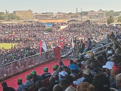 This image depicts a large outdoor public gathering or rally taking place in a stadium or arena under clear, sunny daytime conditions. The setting is consistent with an event in Siaya, Kenya.
The scene is dominated by an immense crowd of people, filling the stadium seats, standing on the field, and extending far into the background. In the immediate foreground, a section of stadium seating is visible, occupied by several individuals mostly with their backs to the viewer, facing a red-carpeted stage or platform. Among these spectators, one person wears a green cap with the text "GENOWA" clearly visible on the back, while another has "CID" partially legible on a dark cap or headband.
On the red-carpeted platform, a central figure, dressed in a red robe or garment, stands as if addressing the crowd. Nearby, other individuals, including security personnel or event staff, are present, along with professional cameras on tripods. Two prominent vertical banners are positioned near the stage; one is red with a white logo and indistinct text (possibly "B O A"), and the other is white with faint dark text (potentially "AKU"). In the distance, above the densely packed crowd, a large video screen displays content.
Beyond the stadium grounds, a cityscape or urban sprawl with various buildings and structures lines the horizon. The overall impression is that of a significant public event, likely a political rally or large community gathering, characterized by a highly engaged and attentive audience.