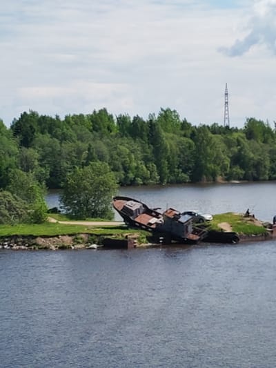 The image shows a partially submerged, decaying ship on a small, grassy island in a body of water, likely a river or lake. The ship appears rusted and dilapidated. On the island, a silver car and what appears to be a black vehicle are also visible. Two individuals are present on the island, one seemingly fishing. In the background is a dense forest and a tall electrical tower. The location is specified as Ivanov Bor, Russia.