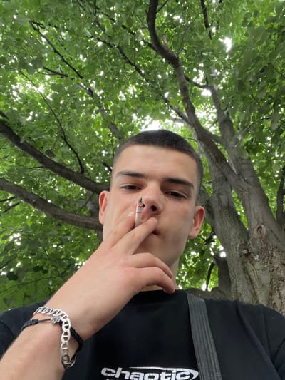 In an outdoor, daytime scene set beneath a canopy of lush green trees, a young man with short hair is captured from the chest up, looking directly into the camera. He holds a lit cigarette between his fingers, bringing it towards his mouth. The sunlight filters through the leaves, creating a dappled effect on his face and the surrounding foliage. He is wearing a black t-shirt, and a silver bracelet with a chain-link design adorns his left wrist. His right arm is bent, with his hand near his face. The backdrop consists of the textured bark of a large tree and the vibrant green leaves of its branches. The overall setting suggests a relaxed, natural environment within the city of Sopot, Bulgaria.