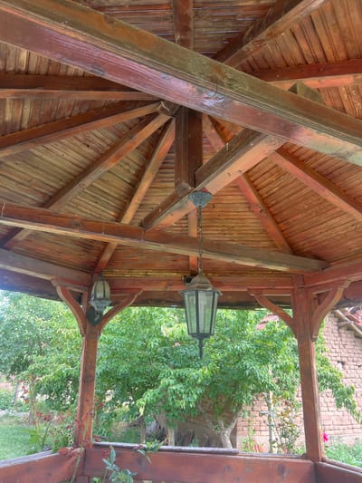 The image shows the interior of a wooden gazebo. The gazebo has a gabled roof made of wooden planks, supported by several wooden beams and struts. Two old-fashioned lanterns hang from the ceiling of the gazebo. One lantern is directly in the center of the image, hanging from a chain, while the other is to the left. The wooden structure appears weathered and aged. Through the open sides of the gazebo, lush green trees and foliage are visible, along with a brick building in the background. The scene depicts an outdoor setting, possibly a garden or a rural area.