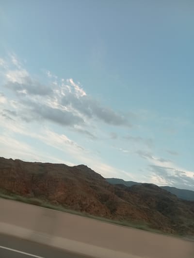 The image depicts a scenic view of a rocky mountain range under a pale blue sky with scattered clouds. The mountains are predominantly brown and tan, with visible texture and ruggedness. They rise steeply from a lower elevation, creating a dramatic silhouette against the sky. A lighter colored structure, possibly a building or a road barrier, is visible on the right side of the frame, running diagonally and obscuring a portion of the mountain. The lighting suggests it is daytime, likely late afternoon or early morning due to the soft, diffused light and the color of the sky. The scene is natural and serene, with no people or specific activities visible. The location, Kok Moynok Eki, Kyrgyzstan, is known for its mountainous terrain and natural beauty, which is consistent with the image. There is no visible text in the image.