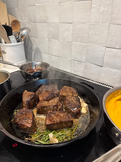 In East Naples, Florida, a close-up shot captures a culinary scene. In the foreground, a cast-iron skillet on a black stovetop holds several thick cuts of seared beef tenderloin. The steaks are glistening, cooked to a medium-rare, and rest in a pool of butter and herbs, including rosemary. A few cloves of garlic are also visible in the pan. To the right of the skillet, a pot filled with a vibrant orange puree, possibly sweet potato or squash, sits on the stove. In the background, a white ceramic crock holds various kitchen utensils, including wooden spoons and a metal whisk, against a light gray tiled backsplash. Another small saucepan with a dark liquid is visible on the stovetop behind the skillet. The image focuses on the preparation of a meal, with the beef steaks being the primary subject.