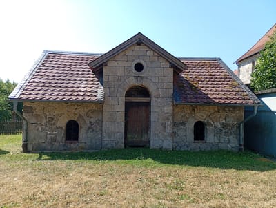 This is a full-shot, eye-level photograph of a small, stone building situated in a grassy area. The building has a gabled roof covered with reddish-brown tiles that appear to be moss-covered in places. The facade is constructed from rough-hewn grey stones. A large, arched wooden door is centrally located, flanked by two small, arched windows with metal grates. Above the arched doorway, there is a circular opening. The building is set against a clear blue sky. The foreground consists of dry, yellowed grass, transitioning to greener grass closer to the building. To the left of the building, a wooden fence is visible. To the right, a portion of another building and some green foliage are present. The building is likely a historical structure, possibly a shed or outbuilding, located in the vicinity of the Walkenried Abbey in Walkenried, Germany. No people or animals are present in the image.