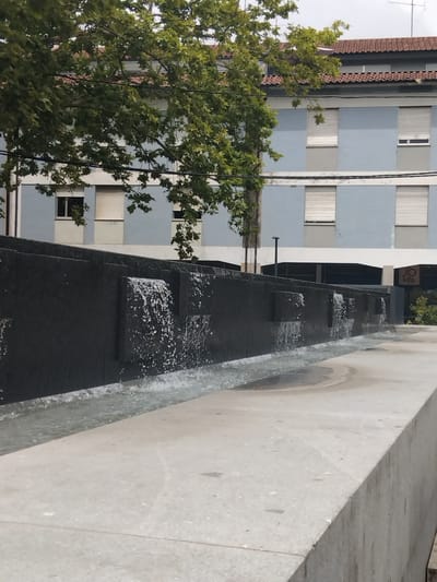 The image depicts an outdoor urban scene in Vila Nova de Famalicão, Portugal. In the foreground, a grey concrete ledge runs diagonally across the frame, with a textured surface. Behind this ledge is a dark, segmented water feature with several sections of cascading water falling into a shallow channel. The water is illuminated, creating a shimmering effect. In the background, a light blue building with horizontal white blinds on its windows and a reddish-brown tiled roof is visible. Green foliage from a tree hangs over the scene from the left. Further in the background, there appears to be a building with signage that reads "JO LTO". The overall impression is that of a public space with a modern water installation.
