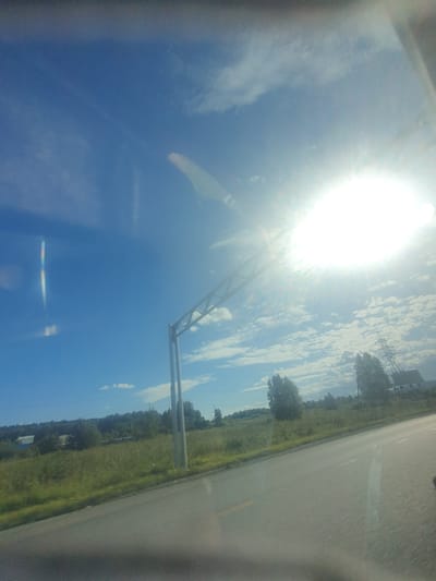 The image captures a bright, sunny day with a clear blue sky and scattered white clouds. The sun is intensely bright, appearing as a white orb in the upper right quadrant of the image, casting a lens flare across the scene. In the foreground, a paved road curves gently to the right, suggesting a highway or a country road. To the left of the road, a grassy field stretches towards a line of trees and vegetation that forms a treeline in the middle ground. In the distance, a few buildings, possibly rural houses or agricultural structures, are visible amidst the trees. To the right of the road, a tall electrical pylon stands prominently against the sky, indicating the presence of power lines in the area. A metal structure, possibly part of an overpass or signage support, is visible in the upper left quadrant of the sky. The overall scene suggests an outdoor environment, likely in a rural or semi-rural setting, with signs of modern infrastructure like roads and power lines. The image appears to be taken from a moving vehicle, as indicated by the slight blur and the angle of the road. While no people are visible, the presence of roads and infrastructure suggests human activity in the area.