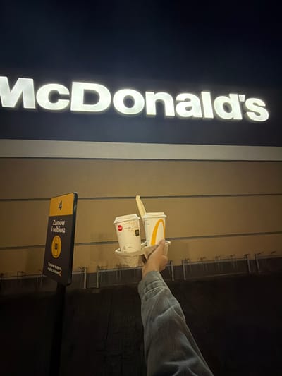 The photograph was taken at night at a McDonald's location in Ustroń, Poland. The McDonald's sign is illuminated on a dark wall. A person's arm, clad in a denim jacket, reaches towards two McDonald's paper cups, presumably to pick them up. The cups are placed on a metal shelf structure, likely a pick-up point for orders. A sign with the number "4" and Polish text "Zamów i odbierz" (Order and Collect) indicates the designated order pick-up location.
