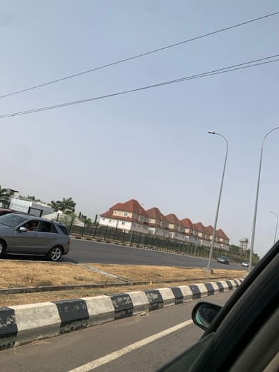 The image presents a roadside scene in Aso, Nigeria, captured from inside a vehicle looking forward and slightly left. The foreground features a black and white striped concrete road barrier, bordering a strip of dry, yellowish-brown grass that serves as a median. Adjacent to the median is a wide, dark grey paved road.

In the midground, on the left side, a silver Mercedes-Benz SUV is either parked or moving slowly on an unpaved shoulder; a person's arm is visible through the open driver's side window. To the right of the main road, two tall, grey streetlights with curved arms extend upwards. In the background, across the main road, a row of approximately seven identical two-story light-colored houses with distinctive orange-red sloped roofs is visible, set back behind a dark fence. Further down the road, two dark-colored sedans are partially visible, and a tall structure, possibly a water tower, stands in the distant background. The sky is hazy and pale blue, with two horizontal power lines visible at the top. Several palm trees are also evident on the far left, behind the SUV.