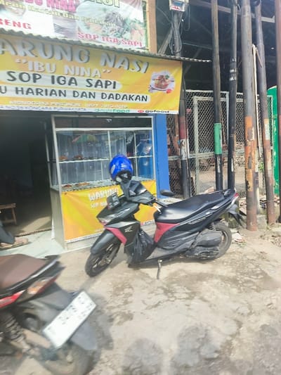 The image shows a street scene in Purwakarta, Indonesia. A black and red Honda Beat scooter is parked in front of a restaurant called "Arrung Nasi 'Ibu Nina' Sop Iga Sapi". The restaurant is situated in a small building with a blue awning. The scooter is parked on the right side of the image, facing the street. The left side of the image shows a portion of a second scooter, parked nearby. The restaurant storefront features a large yellow sign with the restaurant's name in bold lettering, along with a phone number and the word "gofood". The restaurant's window displays a variety of food items on shelves. There are also some wooden posts, a chain-link fence, and a partially visible motorcycle helmet on the left side of the image.