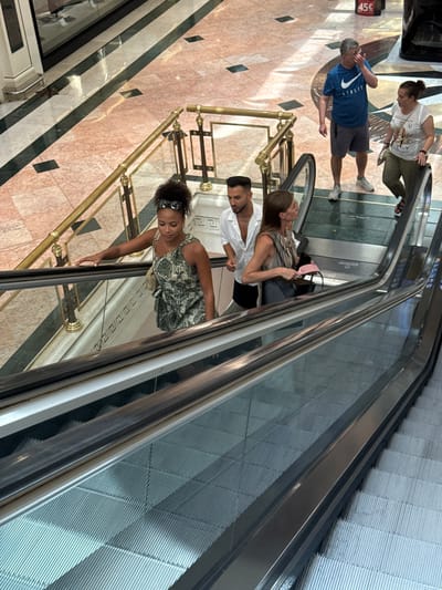 In L'Hospitalet de Llobregat, Spain, a group of people are captured on an escalator within a shopping mall. On the upper level, a man in a blue t-shirt and shorts, and a woman in a white t-shirt and olive pants, are conversing. A price tag displaying "45€" is visible in the background. On the escalator descending, a woman with curly hair and a patterned dress is looking down, while a man in a white open-collared shirt and shorts stands beside a woman in a striped top holding a pink accessory. The escalators are modern, with metal railings and glass panels, set against a backdrop of marble flooring with dark green and black accents.