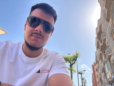 A man wearing sunglasses and a white t-shirt with an Adidas and Marvel logo is taking a selfie outdoors. The background shows a building, trees, and a clear blue sky. The image appears to have been taken on a sunny day in Roquetas de Mar, Spain, as indicated by the architecture and vegetation. The man's face is partially obscured by the sunglasses, but he is looking directly at the camera. The t-shirt has a distinctive Marvel logo in red and white. The building in the background is multi-story with balconies and a light-colored facade, typical of Spanish coastal architecture. The overall scene is casual and bright, suggesting a relaxed outdoor setting.