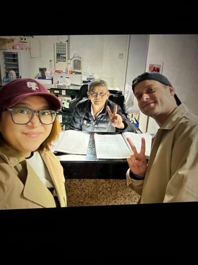 In a dimly lit, cluttered indoor space, possibly a reception area or office, three individuals are captured in a selfie-style photograph. A woman in the foreground on the left, wearing a maroon baseball cap and glasses, smiles warmly at the camera, holding a peace sign with her right hand. Beside her on the right, a man in a dark baseball cap also flashes a peace sign with his left hand, looking towards the camera with a relaxed expression. Between them, an older man wearing glasses and a dark jacket sits at a desk, looking directly at the camera with a neutral expression. The desk is covered with open books or documents. The background reveals shelves filled with various items, including what appear to be papers, boxes, and possibly electronic equipment, suggesting a functional but unorganized environment. The overall scene depicts a casual moment shared by the three individuals in what appears to be a business or service-oriented setting in Taipei, Taiwan.