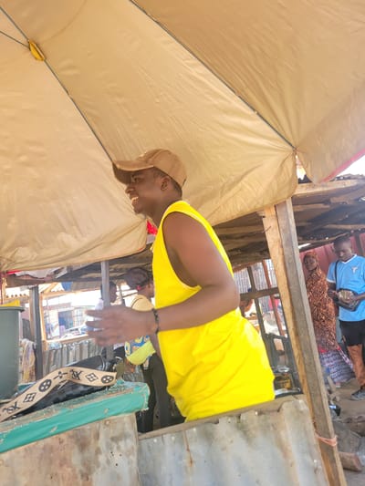 A medium shot captures a young Black man in a tan baseball cap and a bright yellow tank top, smiling and gesturing with his left hand. He stands under a large, tan awning, possibly at an outdoor market. Behind him, to the right, a woman in a patterned dress and headscarf is visible, as is a man in a blue shirt holding a tablet and what appears to be money. The background shows a rustic, wooden market stall structure with glimpses of other people and market goods, suggesting a location in the city of Kwamba, Nigeria. The overall lighting is bright, indicating daytime.