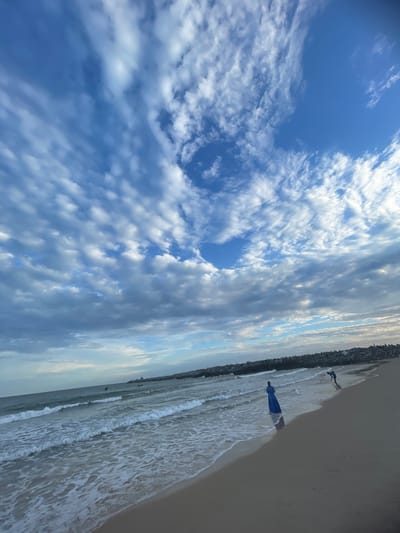 The image shows a beach in Ilado, Nigeria. The sky is blue with scattered white clouds, featuring a circular clear patch. The beach meets the ocean with gentle waves. Two people are visible: one figure further away and another person wearing a blue dress closer to the viewer. A rocky barrier runs along the edge of the beach.