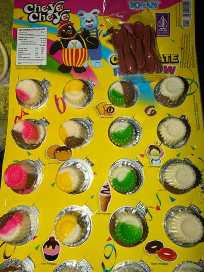 The image shows a yellow package of Choyo Choyo brand jelly candies with a serving suggestion image of ice cream and donuts. The package is arranged in a grid of individual jelly cups. Some of the cups contain jelly in various colors, including pink, white, yellow, green, and brown. There are also small plastic spoons included with the candies. The background is a bright yellow packaging material with cartoon characters of a penguin and a bear. The nutritional information and ingredients are listed in Indonesian on the package. The product appears to be a snack food. There is no discernible location in East Jakarta, Indonesia.