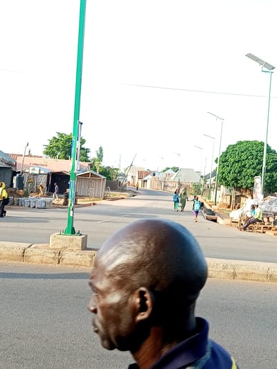 The image captures a street scene in Tumfure, Nigeria, during what appears to be daytime with a bright, overcast sky. In the foreground, a man's head and shoulders are visible in profile, facing to the left. His dark skin is glossy, suggesting sweat or oil, and his head is bald and shiny. He is wearing a dark blue polo shirt with a lighter blue collar and a yellow stripe on the sleeve.
The background depicts a street with several people walking. To the left, a person in a yellow top and black pants is walking away from the camera. Further down the street, there are shops and structures made of corrugated metal and wood, some with open fronts. A prominent green pole, possibly a lamppost or utility pole, stands near the center of the image, with several flyers or posters attached to it.
Further down the street, a group of children and adults are walking. One child wears a blue backpack, and another is dressed in a light-colored garment. A large, leafy green tree stands on the right side of the street, providing shade to some of the structures. In the distance, more buildings and trees are visible, and several modern streetlights with solar panels line the road. On the right side of the road, two men are seated on a bench outside a shop. One man wears a lime green shirt and is looking towards the street. The other man, in a blue shirt, is looking down.
The overall atmosphere suggests a normal day in a town or city. There are no overt signs of distress or unusual activity. The presence of modern streetlights indicates some level of development. No visible text is legible in the image.