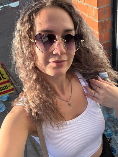 The image shows a close-up, rotated view of a young woman with long, curly blonde hair. She is wearing a white ribbed tank top and a delicate silver necklace with a small cross pendant. Her face is partially visible, and she is wearing large, heart-shaped sunglasses with dark lenses. She has several rings on her fingers, including one with a small stone.
In the background, a brick wall can be seen to the left. To the upper right, several plastic water bottles are visible. The lighting suggests it is daytime, likely sunny given the shadows and highlights on her skin and hair. The overall setting appears to be outdoors, possibly in an urban or park-like environment, as indicated by the brickwork and the hint of a street scene with some signage at the bottom.
There is some text visible on a yellow and red flag-like object at the bottom of the image, but it is partially obscured and difficult to read clearly. The orientation of the image is rotated, making the subjects appear sideways. The woman's pose and expression, though not fully visible, suggest a casual and relaxed demeanor.
