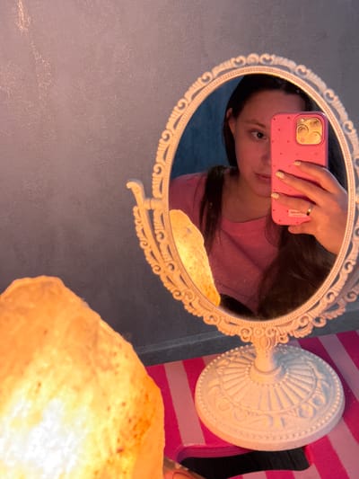This media file captures a close-up reflection in an ornate, oval mirror with a white, scrolled frame. The reflection shows a young person holding a pink cell phone with a polka-dot case. The phone's camera is visible, indicating they are taking a selfie. The person is wearing a pink t-shirt and has a ring on their finger. Their gaze is directed at the camera. The background of the reflection is dark, with hints of brown hair.
To the right of the mirror is a white, decorative base that supports the mirror. This base has intricate embossed patterns. Behind the mirror and its stand, a pink and white striped surface is visible, suggesting a patterned background or furniture. In the foreground, out of focus, is a glowing, translucent object, possibly a salt lamp, emitting a warm, yellow light.
The lighting suggests it might be evening or indoors with artificial light, given the warm glow. The overall mood appears casual and personal, typical of a selfie. There is no visible text in the image. The location context provided is Prikubanskiy, Russia, but there are no specific visual cues within the image to confirm this location.