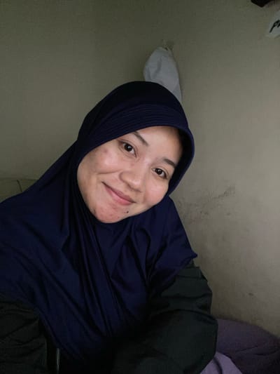 A selfie captures a woman wearing a dark blue hijab and dark clothing. She is smiling and looking directly at the camera. Her face shows some texture and minor blemishes. The background is a plain, textured wall with subtle shadows. The overall lighting is dim, suggesting an indoor environment, possibly a room in a residential building in Banjarbaru, Indonesia, given the style of the hijab and the typical interior finishes seen in the region. There is a white object, possibly a plastic bag, hanging on the wall behind her.