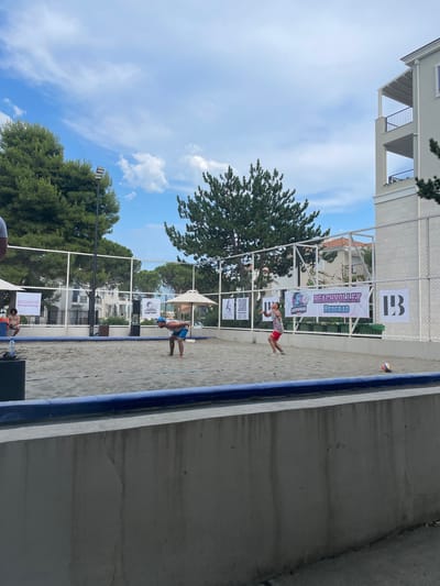 In Radovići, Montenegro, a beach volleyball court is set up outdoors under a cloudy sky. Two men are actively engaged in a game on the sandy court, with one man in the foreground in a blue cap and shorts, bending low in a defensive stance, and another man in a red and white shirt and red shorts on the opposite side of the net, in the process of serving or hitting the ball. A volleyball is visible in the air near the second man, and another volleyball rests on the sand near the side of the court. The court is surrounded by a white net fence, and banners advertising "BEACHVOLLEY BEOGRAD" are displayed behind the net. Several large trees are visible behind the fence, and a multi-story building with balconies is located to the right of the court. The court itself is bordered by blue padding.