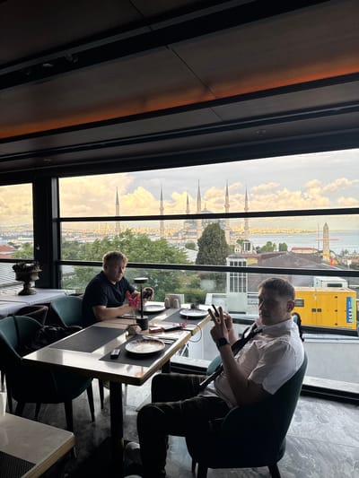 Two men are seated at a table in a restaurant with a large window overlooking the city of Istanbul, Turkey. The man on the left is wearing a black t-shirt and appears to be looking at his phone. The man on the right is wearing a white collared shirt and a tie, and is holding up two fingers in a peace sign gesture while looking at his phone. The table is set with plates, cutlery, and a small vase with flowers. In the background, the Sultan Ahmed Mosque (Blue Mosque) and other minarets are visible against a cloudy sky. Beyond the mosque, the Bosphorus strait and a yellow generator are visible. The restaurant appears to have modern decor with dark wood paneling and upholstered chairs.